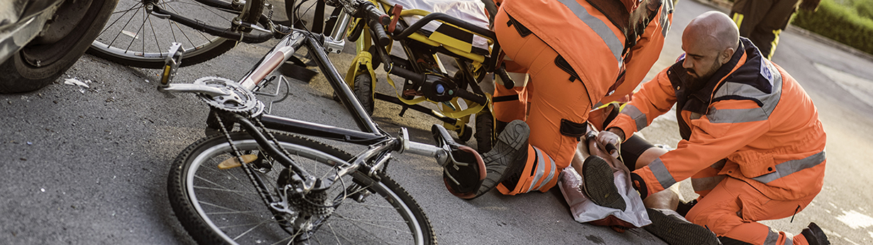 Paramedic team knealing around injured cyclist lying on street, damaged bicycle and car beside them, firefighters and fire engine in background.