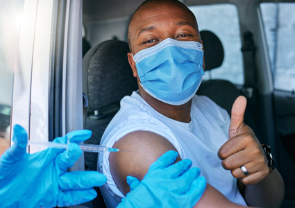 Drive thru covid and corona virus vaccine site as a public service for the people. Man giving thumbs up and endorsing the jab while wearing a mask to avoid infection, approving being vaccinated