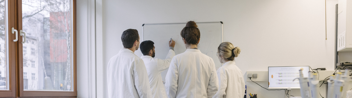 A group of medical students discussing some work while huddled around a whiteboard at the lab together.