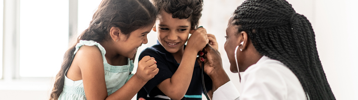 A brother and sister duo of Indian decent, visit the doctor together.  They are sitting up on the exam table and playing with the doctors stethoscope.   The young by is being silly and holding the end of the stethoscope up to his ear, along with the help of the female African doctor, who is bent down in front of them. The young girl is giggling as her brother plays pretend.